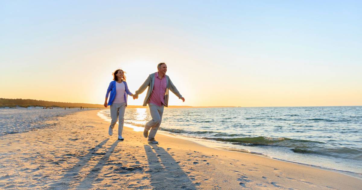 een oudere man en vrouw die over het strand rennen met de zon achter zich
